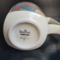 Kaffeebecher, eckig-rund, cremeweiß matt, mit bunten Motiven an den Außenseiten. Unterseite mit Stempel