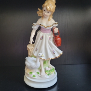 Mädchen mit geblümtem Kleid und Goldrändern. An ihrer Seite eine Gans, am linken Arm einen roten Wasserkübel tragend